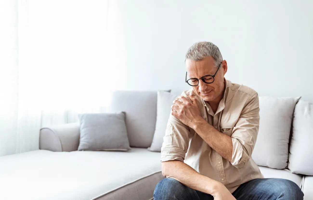 A middle-aged man sits on a couch, holding his shoulder in pain, representing common conditions treated by platelet-rich plasma (PRP) therapy.