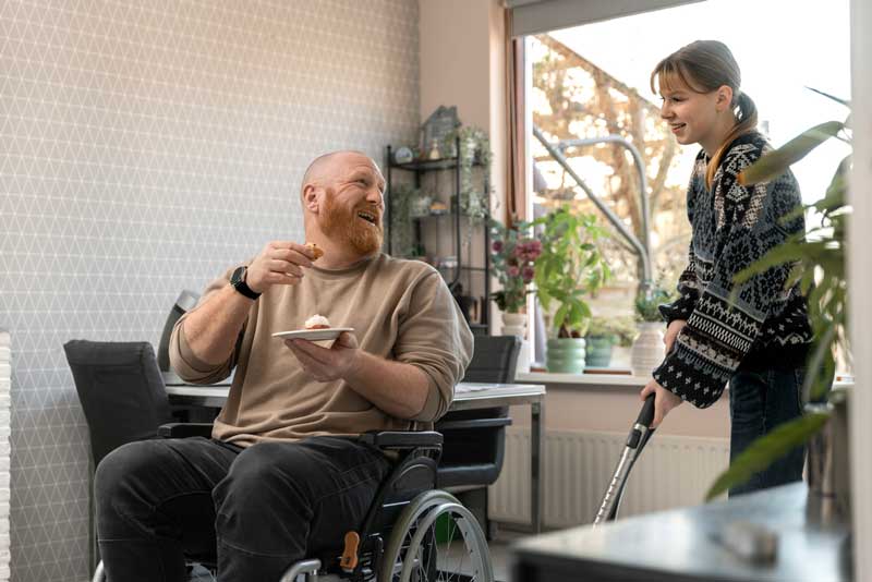 A smiling man with ALS sitting in a wheelchair enjoys a piece of cake while his caregiver vacuums the living room, highlighting daily support and quality of life.