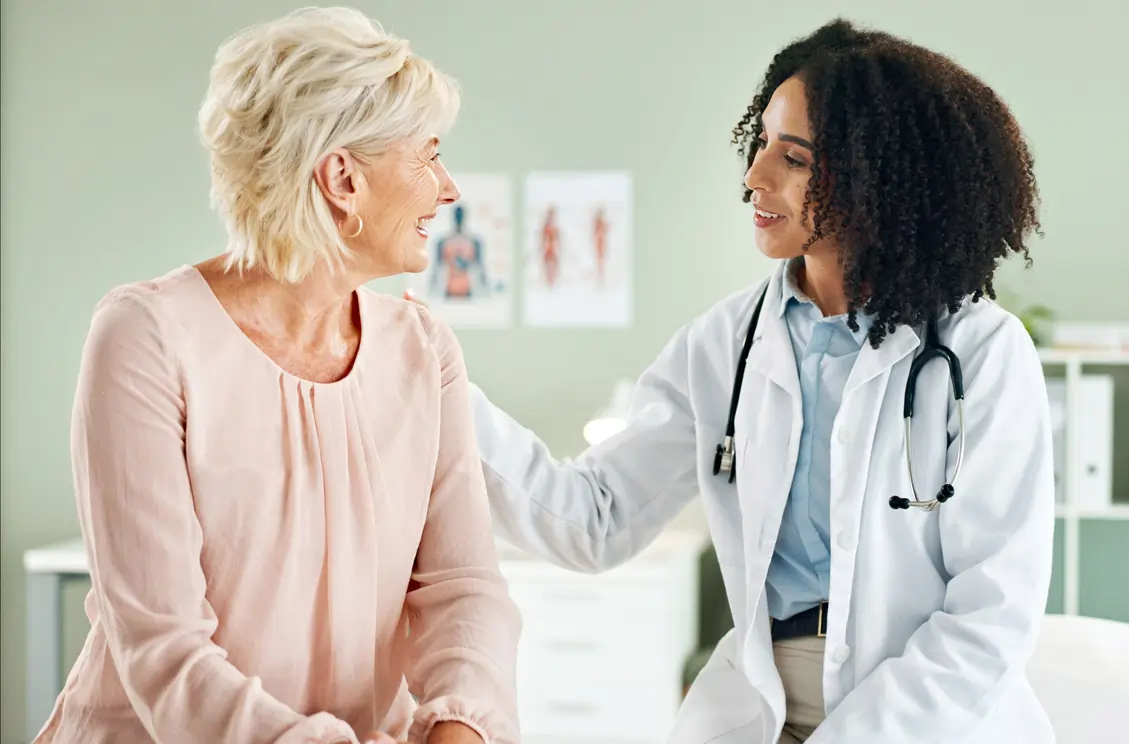 A female doctor in a white lab coat with a stethoscope comforting a smiling senior woman sitting in a bright medical office, representing the trusted regenerative medicine and mesenchymal stem cell therapy offered at the R3 Stem Cell Puerto Vallarta treatment clinic.